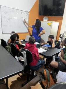 Sala com estudantes em mesas e uma educadora escrevendo no quadro