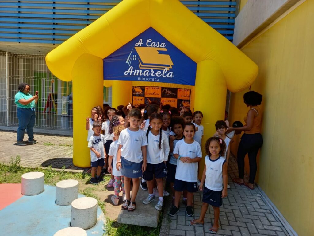 Crianças em frente à biblioteca inflável amarela da Casa Amarela, com a placa do projeto ao fundo